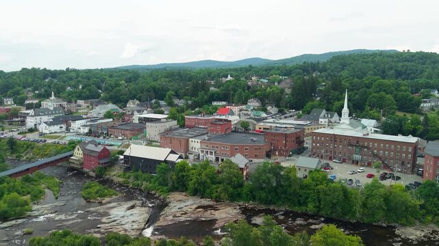 Littleton, New Hampshire, high aerial drone video over the Ammonoosuc River, showing the historic covered bridge, red grist mill, and downtown village architecture set against forested green hills.