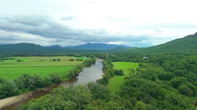 Groveton, New Hampshire, high aerial drone video over lush green forests, farmland, and a winding river bend, set against distant mountains under a dark, dramatic stormy sky.