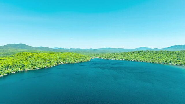 Maidstone Lake, Vermont, aerial hyperlapse motion shot gliding over deep blue waters toward lush green forested shorelines and distant mountain peaks under a clear summer sky.