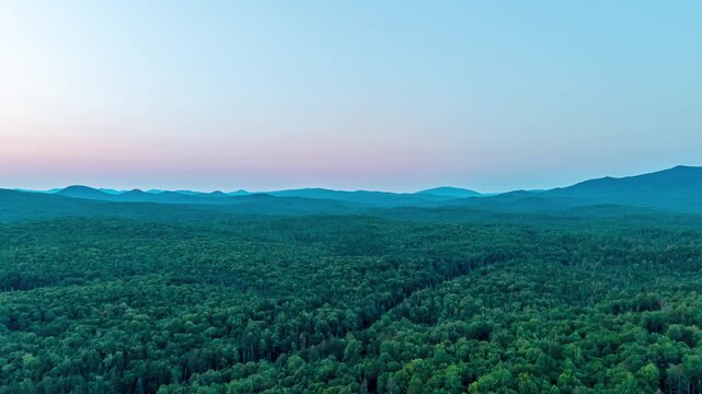 Northern Vermont aerial hyperlapse video showing a vast green forest canopy stretching toward distant mountain ridges during a soft pastel dusk sunset.