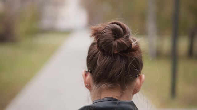 Close up of woman with brown hair in bun walking away on outdoor path. Her black leather jacket and multiple ear piercings are visible as woman moves through soft focus park setting