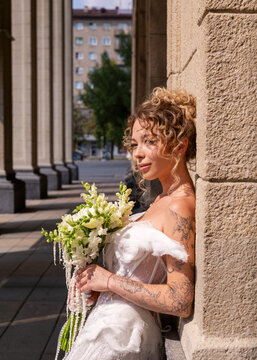Tattooed bride with bouquet leaning against stone column outdoors