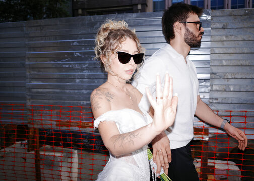 Bride and groom in sunglasses standing outdoors near fence