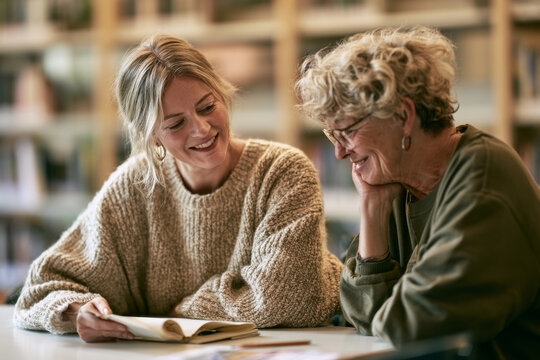 A woman in her fifties reads aloud from a notebook beside a volunteer tutor at a bright library table during an adult class. Adult education literacy teaching how to read background with copy space.