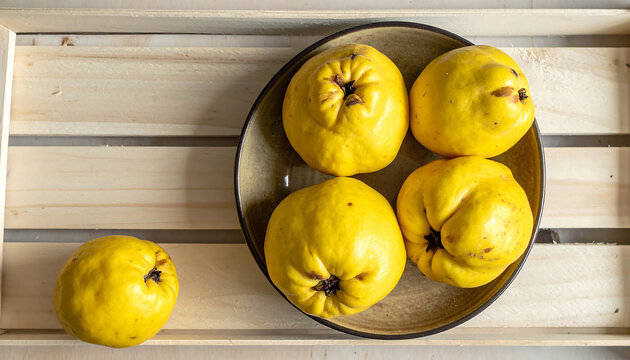 Four bright yellow quince fruits, three inside a ceramic bowl and one outside, placed on a light wooden crate surface with natural lighting