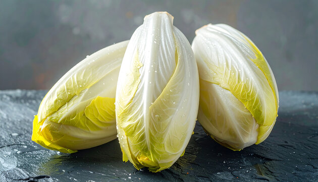 Three fresh heads of belgian endive with pale yellow and white leaves arranged on a dark textured slate surface with soft lighting and water drops