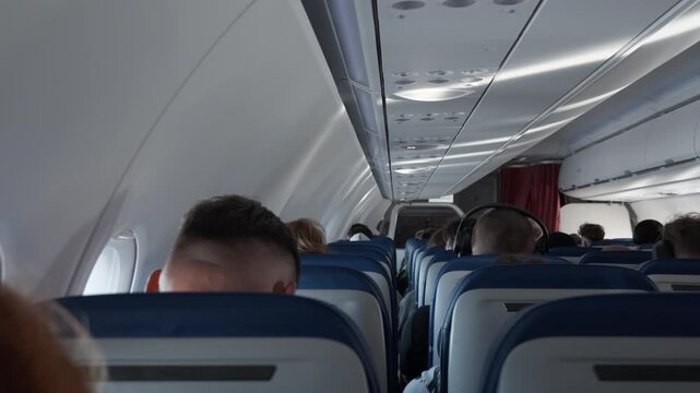 Passengers sit in seats inside commercial airplane cabin during flight. People travel by passenger aircraft on vacation or business trip. Airline transport interior with overhead luggage bins.
