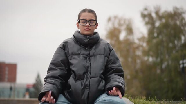 Close up of young woman meditating outdoors on stone steps wearing black puffer jacket and blue jeans. Hands rest gently in lap with soft lighting under overcast sky. Trees and urban