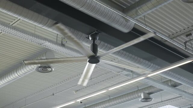 Large industrial ceiling fan rotates under modern building roof structure. Ventilation duct pipes and climate systems circulate cool air in warehouse. Engineering and HVAC cooling concept.
