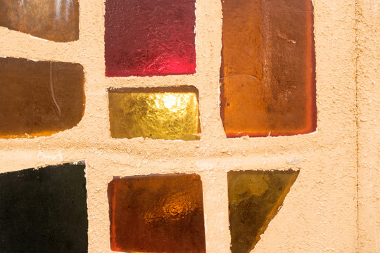 Abstract close up of colorful stained glass squares set in a rough white plaster wall, warm red amber and yellow tones glowing in sunlight, minimal geometric architectural detail, grainy film look