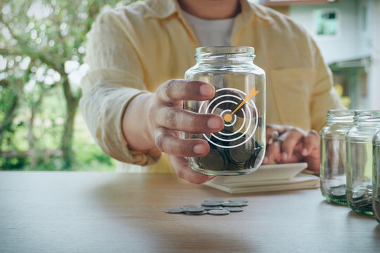 Person holding glass savings jar filled with coins featuring a target and dart hologram icon, concept of financial goal achievement, money saving target, success, and investment planning.