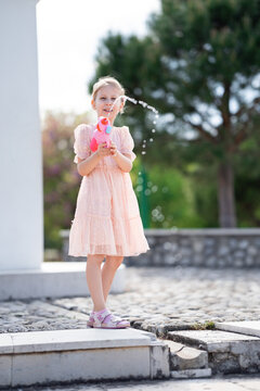 Summer park scenery featuring joyful child splashing water gun standing on stone pavement wearing light pink summer dress with bokeh greenery and warm afternoon glow in full body composition