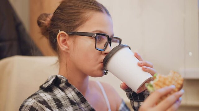 Close up of young woman drinking from white coffee cup while holding and eating food in indoor setting. Woman wears glasses has her hair tied back with visible earrings and is dressed