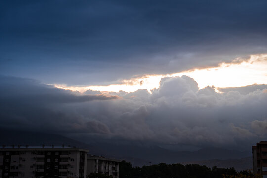 Stormy clouds illuminated by the setting sun casting golden rays through the thick grey layers above the residential blocks creating a dramatic sky filled with light and shadow textures