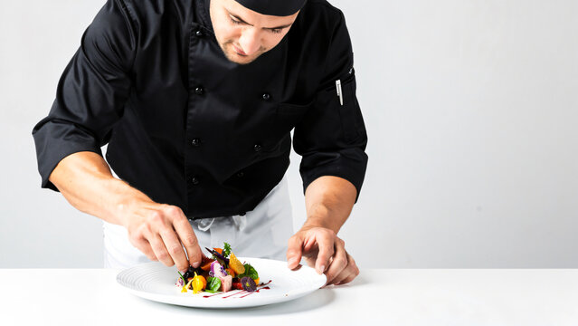 Professional chef in clean black uniform carefully plating gourmet dish in modern kitchen studio, precise hand movement and fine dining presentation, elegant food styling with microgreens .