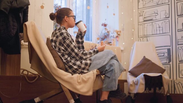 Young woman with glasses sits in cozy indoor setting drinking from white cup and holding food while wearing casual attire. scene features warm lighting decorative elements like string lights