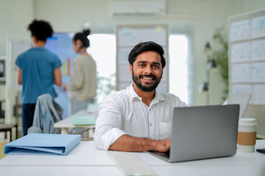Smiling businessman holding a coffee cup at his desk, looking confidently at the camera with colleagues working in the background of a bright office.