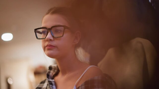 Close up of young woman with glasses and plaid shirt softly lit in warm indoor setting. Woman remains still wearing casual clothing including visible tank top straps under open collar