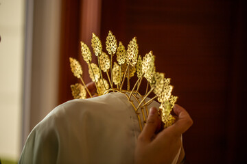 Rear view of hands carefully adjusting an intricate gold filigree tiara crown onto a bride wearing a white hijab veil. © udeyismail