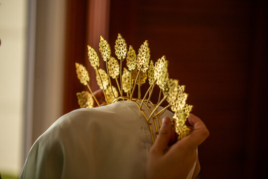 Rear view of hands carefully adjusting an intricate gold filigree tiara crown onto a bride wearing a white hijab veil.