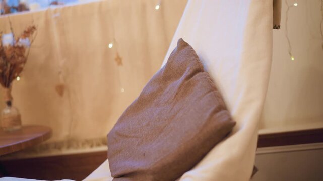 Close up of brown fabric pillow resting on off white chair with soft lighting and blurred background. scene features warm ambient light decorative string lights in distance wooden table