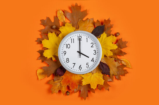Round clock set to 4 o'clock surrounded by autumn leaves on orange background. Flat lay for daylight saving time change, fall season sales, early morning, end of workday, time management. Top view.