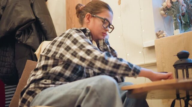 Close up of young woman wearing glasses and casual clothing seated at wooden table in indoor setting. Woman interacts with objects on tabletop including adjusting or moving them while soft