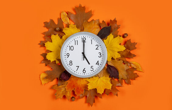 Top view of round analog clock set to 5 o'clock surrounded by fall leaves on orange background. Flat lay for Daylight Saving Time, autumn sales, end of workday, or time management concepts.