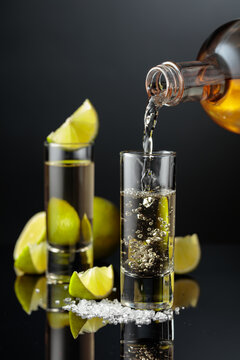 Tequila shots with salt and lime slices on a black reflective background.