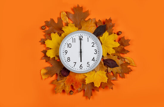 Round analog clock set to 6 o'clock surrounded by fall leaves on orange background. Flat lay composition for daylight saving time, autumn sales, end of workday, evening time, or time management. 