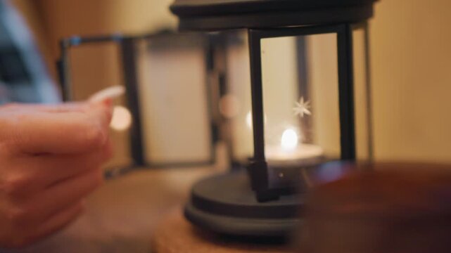 Close up of hand with long nails holding and releasing unlit tealight candle in front of lit black metal lantern. soft glow illuminates scene revealing warm indoor lighting and blurred