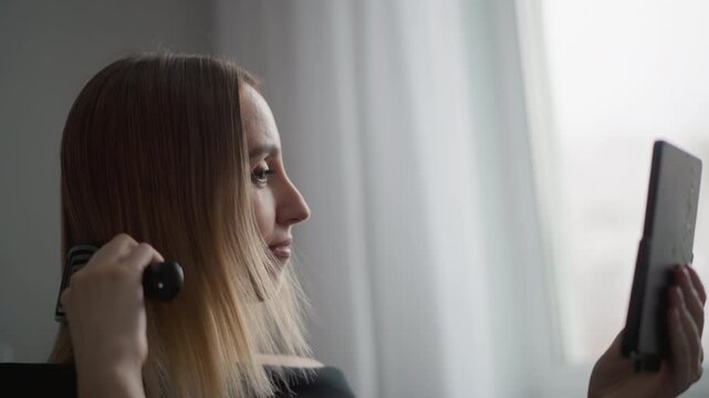 Close up of woman brushing her hair while looking at herself in mirror. Woman holds black comb and tablet woman is using to check her reflection with soft natural light coming through white