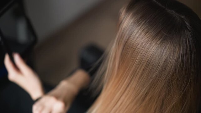 Close up of woman brushing long straight hair with smooth texture while looking into handheld mirror. subject holds brush in one hand and uses another to steady her position indoors