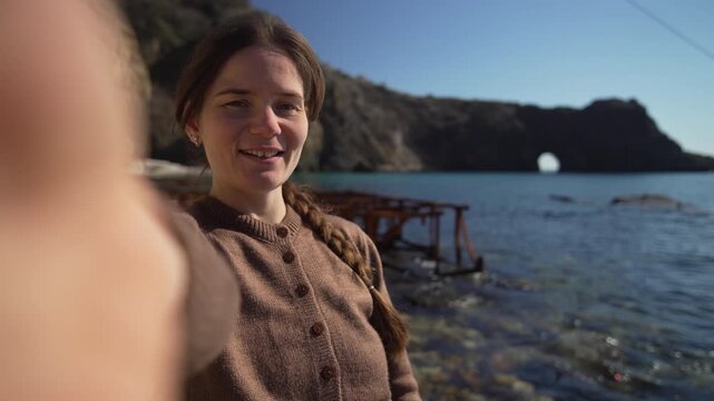 Woman beach selfie standing on rocky shore waving and showing natural rock arch in the sea during a sunny travel vlog