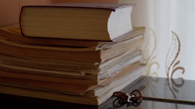 Old, tattered books stacked on a reflective surface next to a window, symbolizing the intersection of faith, knowledge, education, and history