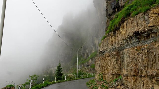 Dramatic view of an empty mountain pass winding alongside a sheer rock face on a moody and rainy day. Thick, low clouds and mist envelop the epic landscape creating a mysterious atmosphere