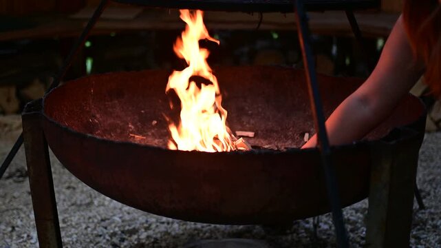 Video footage of bright orange flames flickering inside a large metal fire pit bowl suspended on a tripod during a calm evening.