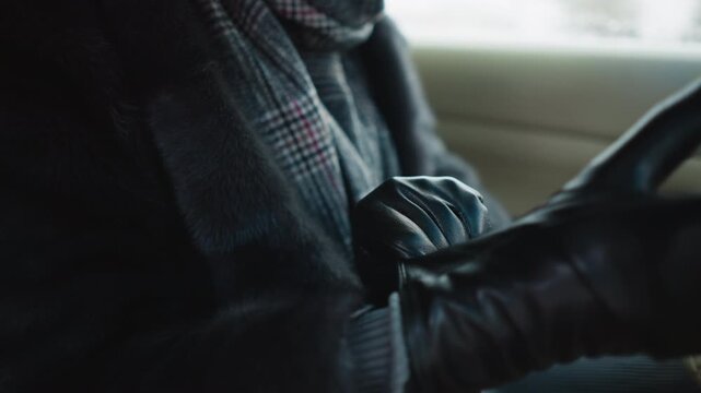Close up of woman putting on black leather gloves while wearing dark coat and plaid scarf inside vehicle. scene shows texture detail of gloved hands against soft indoor lighting with natural
