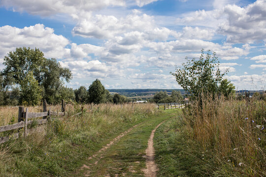 Rural landscape with a dirt path leading through a meadow toward distant fields and trees under a dramatic cloudy sky.