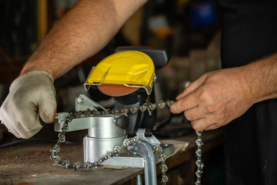 A master is sharpening a chainsaw chain using a machine. The workshop is busy with tools and equipment. It is daytime and the place is well-lit