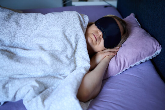 Woman sleeping in a bed covered with a blanket while wearing a black sleep mask on her eyes to block light.