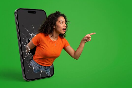 Studio portrait of young woman in orange t-shirt bursting through shattered smartphone screen and pointing, isolated on green chroma key background