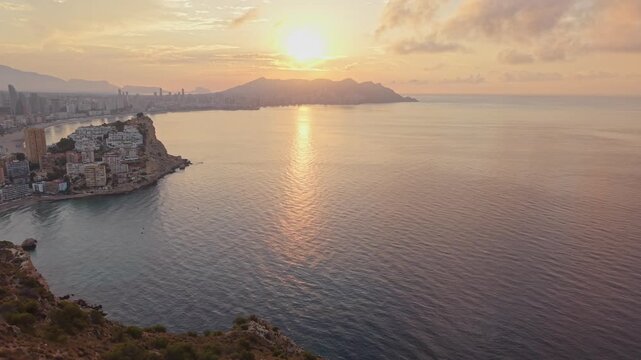 The sun is setting over the water in Altea. The sky shows colors as light reflects off the sea. The city and mountains are visible in the background during the evening.