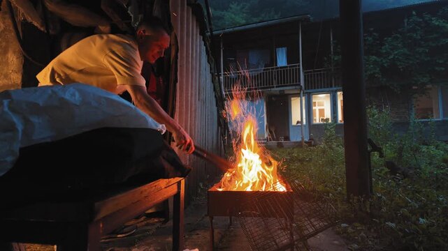 Caucasian man stirring burning wood with tongs in a barbecue grill, preparing for a cookout in his backyard at night with sparks flying from the bright orange flames against a dark background