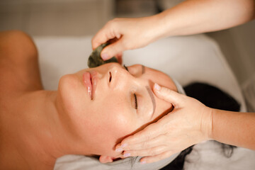 Professional masseuse performing gua sha facial massage with jade stone tool in spa © cenchild