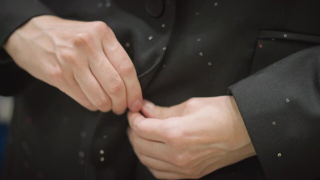 Close up of hands fastening black jacket with small reflective dots. person manipulates fabric and buttons demonstrating precise manual dexterity against soft lighting. Background remains