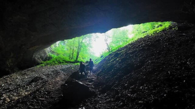 Two adventurous tourists explore the dark Sakinule Cave in Georgia, climbing a steep rocky path to a bright, sun-drenched entrance, discovering a beautiful green forest on a sunny day