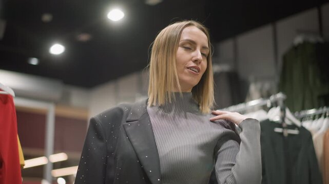 Close up of woman examining clothing in store adjusting her jacket and looking around. Woman wears sparkly blazer over ribbed turtleneck with various garments on racks behind her. Soft