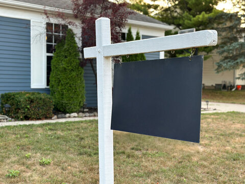 A blank real estate sign hangs in front of a suburban home, surrounded by a landscaped lawn, driveway, and garage. Ideal for property, housing, real estate, and neighborhood concepts.