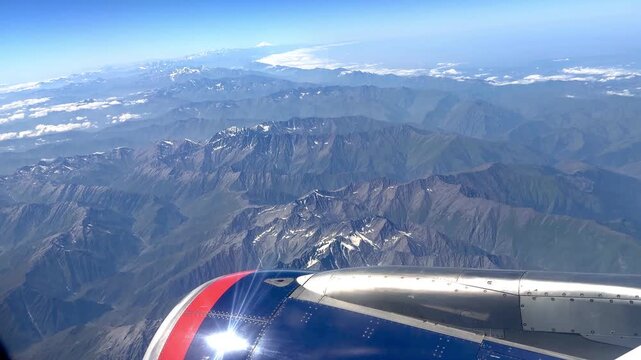 Aerial perspective of a mountain range with visible airplane wing, highlighting snow-covered peaks and expansive blue sky above